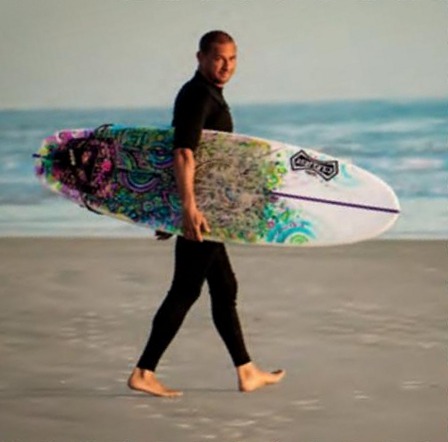 Jorge Alvarado in a black wetsuit walking barefoot along the beach with a colorful surfboard under his arm, with the ocean behind him