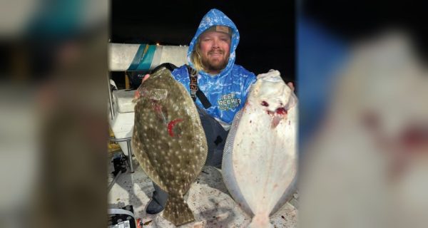 Capt. John Ellis in a blue Reef Wrecker hoodie holding up two big flounder on his boat at night, one dark side up and one white side up