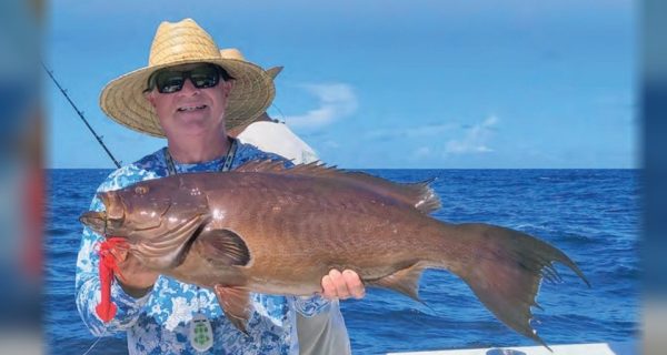 An angler in a straw hat, sunglasses and a blue and white patterned shirt holding up a big grouper with a red jig in its mouth on a boat offshore