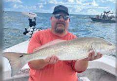 An angler in a cap, sunglasses and an orange shirt sitting on a boat holding a redfish, with a trolling motor, another boat and puffy white clouds behind him