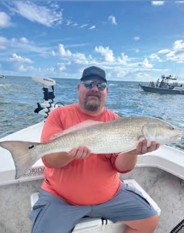 An angler in a cap, sunglasses and an orange shirt sitting on a boat holding a redfish, with a trolling motor, another boat and puffy white clouds behind him