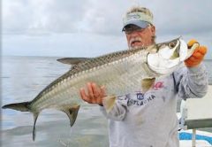 An angler in a visor, sunglasses and orange gloves holding up a big tarpon on a boat over the shallow flats, under a cloudy sky
