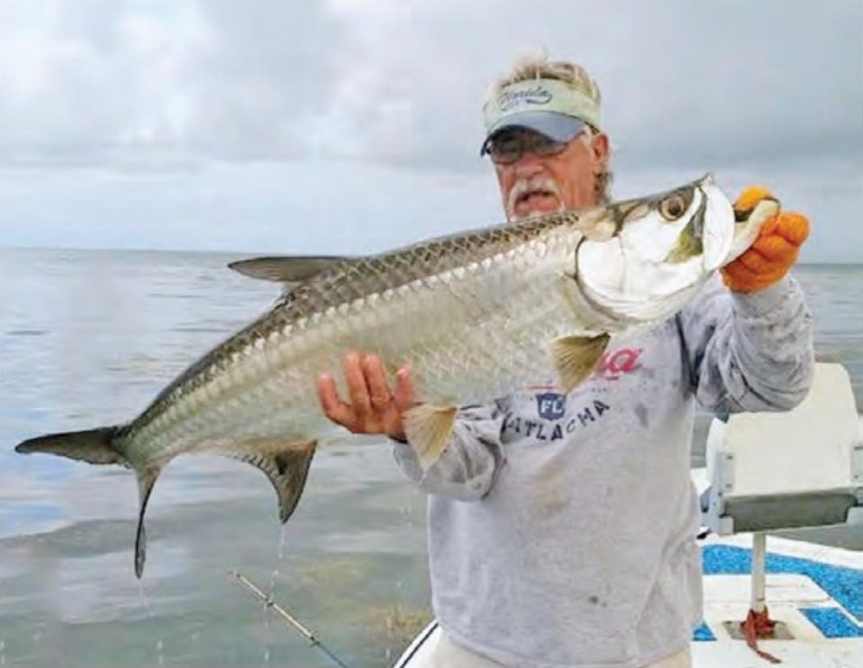 An angler in a visor, sunglasses and orange gloves holding up a big tarpon on a boat over the shallow flats, under a cloudy sky