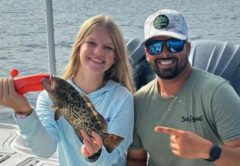 Youth angler Azalea smiling on a boat as she holds up her first grouper with an orange fish grip, while Capt. Evan Sanders in a white cap and sunglasses points to her catch