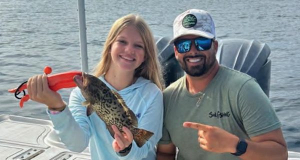 Youth angler Azalea smiling on a boat as she holds up her first grouper with an orange fish grip, while Capt. Evan Sanders in a white cap and sunglasses points to her catch