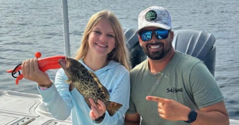 Youth angler Azalea smiling on a boat as she holds up her first grouper with an orange fish grip, while Capt. Evan Sanders in a white cap and sunglasses points to her catch