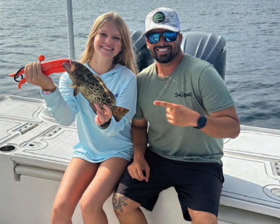 Youth angler Azalea smiling on a boat as she holds up her first grouper with an orange fish grip, while Capt. Evan Sanders in a white cap and sunglasses points to her catch
