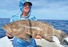 An angler in a blue hooded sun shirt and cap holding up a big grouper on a boat, with the open ocean and blue sky behind him