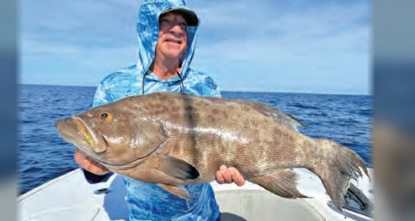 An angler in a blue hooded sun shirt and cap holding up a big grouper on a boat, with the open ocean and blue sky behind him