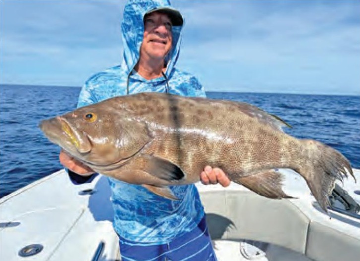 An angler in a blue hooded sun shirt and cap holding up a big grouper on a boat, with the open ocean and blue sky behind him