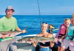 A family on a charter boat with their catch: a man in a cap and green shirt and a smiling woman each holding a kingfish, beside two smiling girls in life jackets, with the open ocean behind them