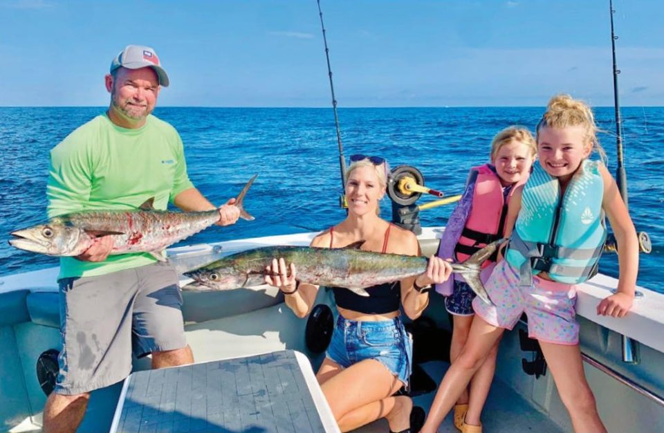 A family on a charter boat with their catch: a man in a cap and green shirt and a smiling woman each holding a kingfish, beside two smiling girls in life jackets, with the open ocean behind them