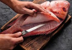 Hands filleting a red snapper with a fillet knife on a dark wooden cutting board