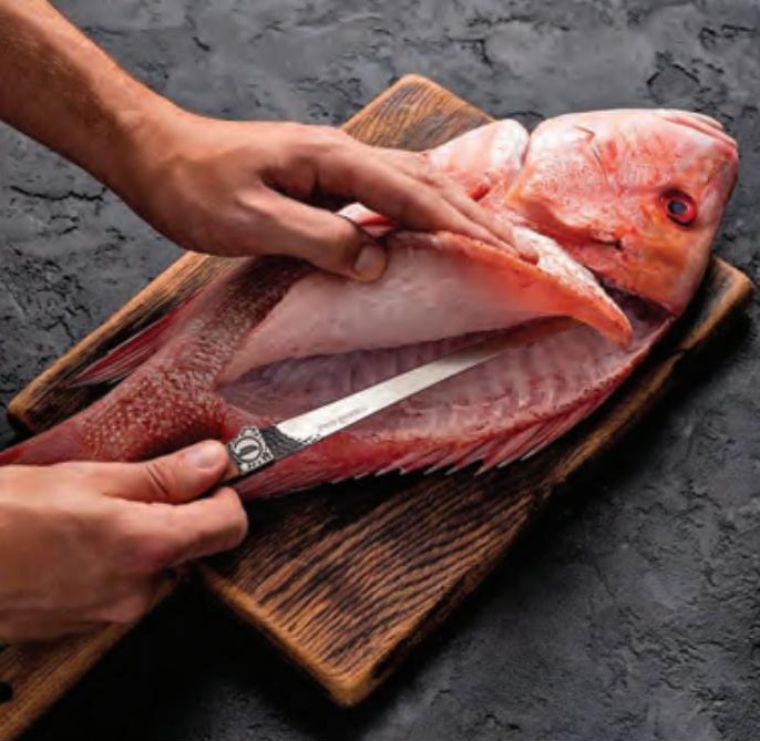 Hands filleting a red snapper with a fillet knife on a dark wooden cutting board