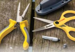 Yellow and gray fishing pliers and yellow Smith's fishing scissors on a weathered wooden dock, beside a gray sheath, a white jig and a sinker on a line