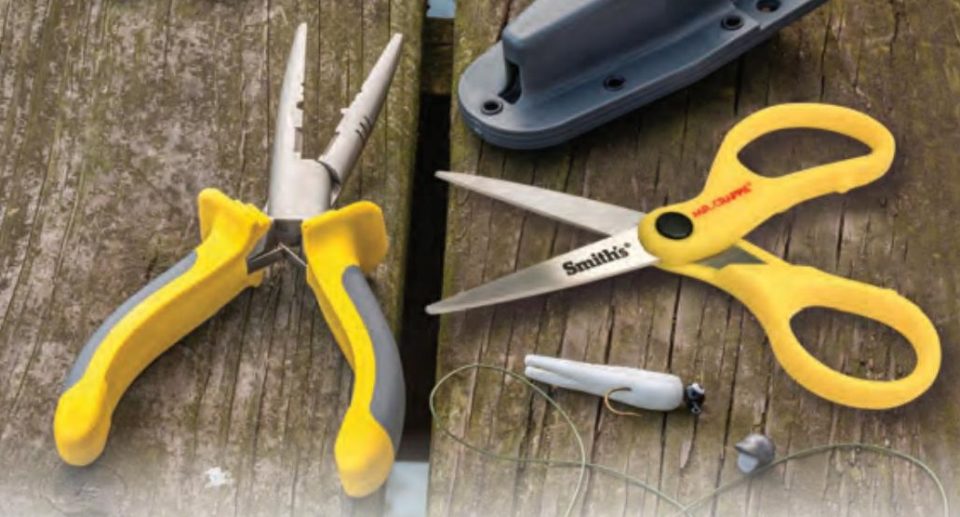 Yellow and gray fishing pliers and yellow Smith's fishing scissors on a weathered wooden dock, beside a gray sheath, a white jig and a sinker on a line