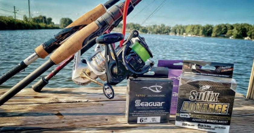 Spinning rods and reels, one spooled with green braid, on a wooden dock beside boxes of Seaguar fluorocarbon and Sufix Advance monofilament line, with a lake and trees behind them