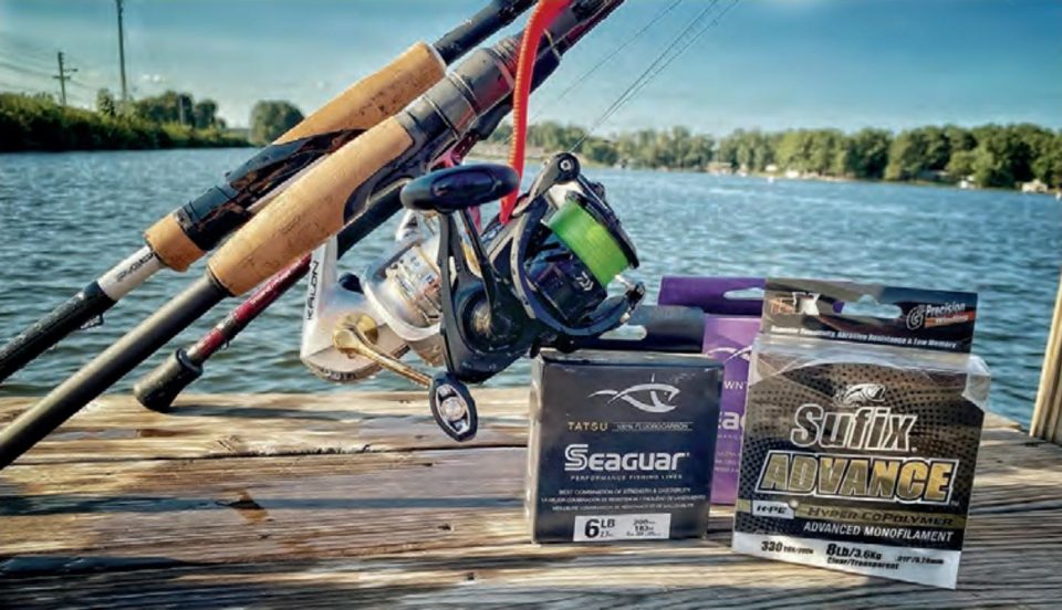 Spinning rods and reels, one spooled with green braid, on a wooden dock beside boxes of Seaguar fluorocarbon and Sufix Advance monofilament line, with a lake and trees behind them