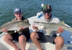 A smiling boy in a cap holding a redfish and a man in a camo cap and sunglasses holding a snook, sitting side by side on the bow of a boat