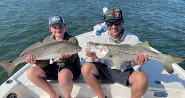 A smiling boy in a cap holding a redfish and a man in a camo cap and sunglasses holding a snook, sitting side by side on the bow of a boat