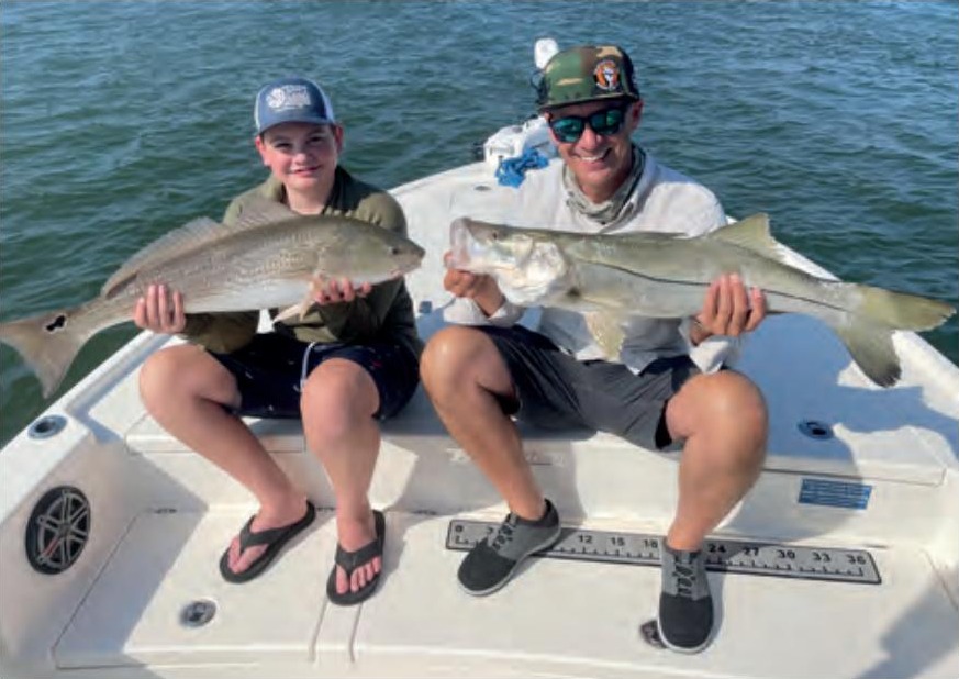 A smiling boy in a cap holding a redfish and a man in a camo cap and sunglasses holding a snook, sitting side by side on the bow of a boat