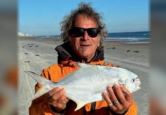 Marco Pompano in sunglasses and an orange jacket holding a pompano on the beach, with the ocean behind him
