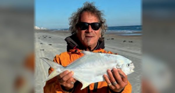 Marco Pompano in sunglasses and an orange jacket holding a pompano on the beach, with the ocean behind him