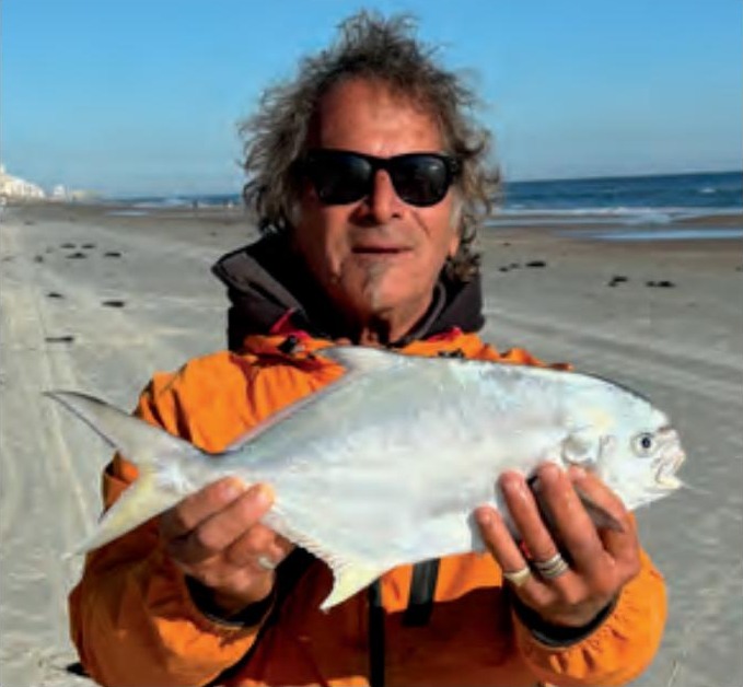 Marco Pompano in sunglasses and an orange jacket holding a pompano on the beach, with the ocean behind him