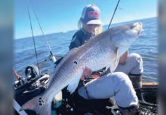 Wendy Biles in a cap holding a giant bull redfish in her kayak, with fishing rods beside her and the ocean beyond