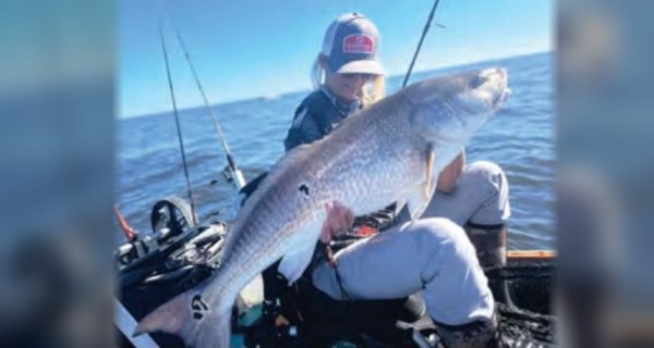 Wendy Biles in a cap holding a giant bull redfish in her kayak, with fishing rods beside her and the ocean beyond