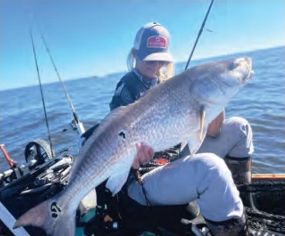 Wendy Biles in a cap holding a giant bull redfish in her kayak, with fishing rods beside her and the ocean beyond