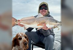Capt. Will Wolfson in a hood and sunglasses holding up a redfish on his boat, with his orange and white dog sitting in front of him