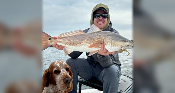 Capt. Will Wolfson in a hood and sunglasses holding up a redfish on his boat, with his orange and white dog sitting in front of him