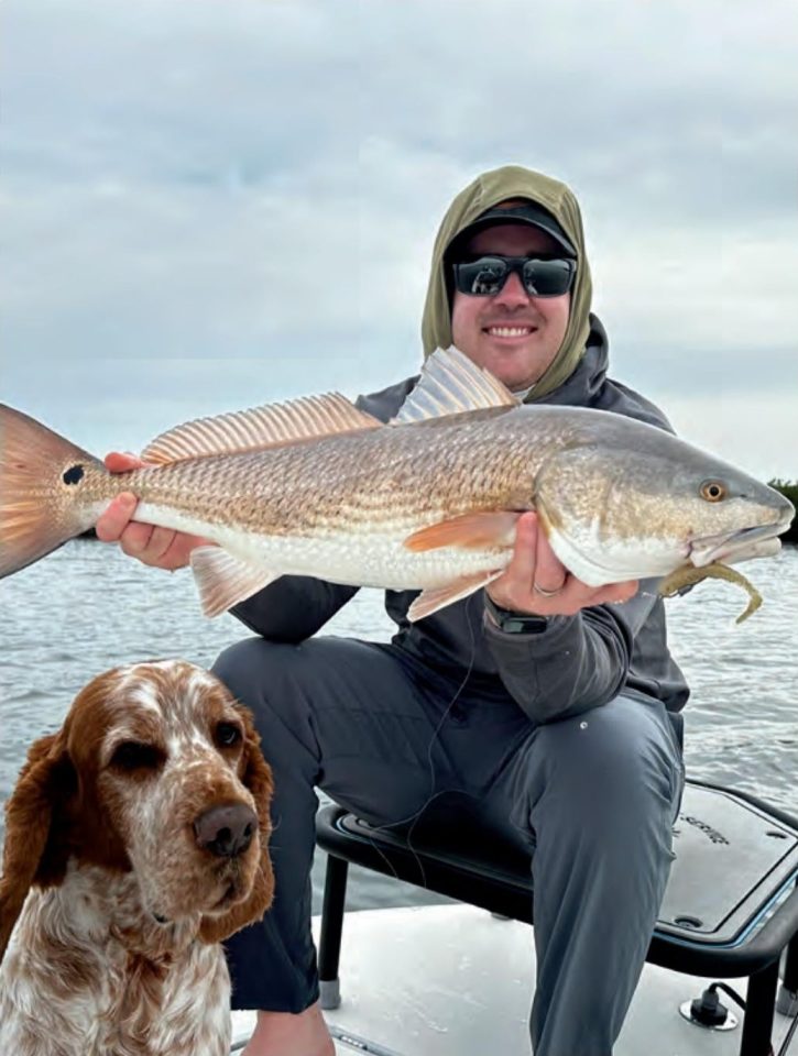 Capt. Will Wolfson in a hood and sunglasses holding up a redfish on his boat, with his orange and white dog sitting in front of him