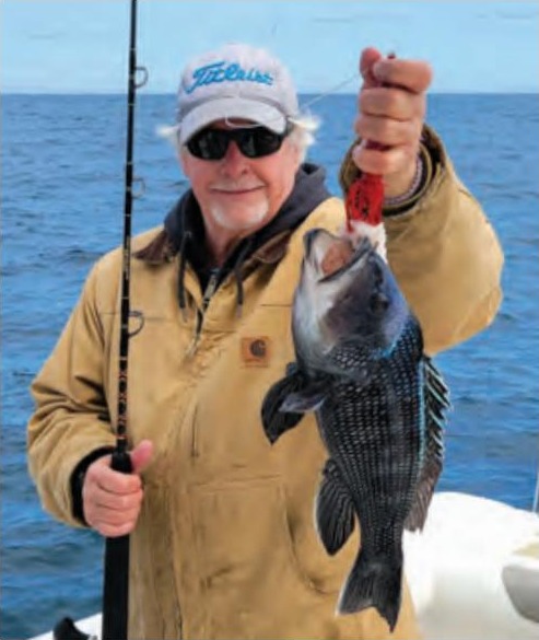 A smiling angler in a white cap, sunglasses and a tan work jacket holding a rod in one hand and a black sea bass on a jig in the other, with the ocean behind him