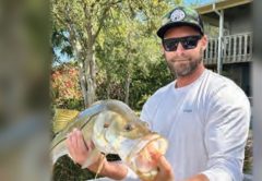 Capt. Brian Revak in a camo cap and sunglasses holding up a snook in front of a house and trees