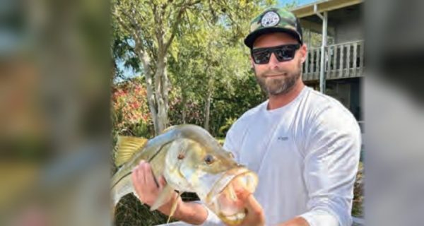 Capt. Brian Revak in a camo cap and sunglasses holding up a snook in front of a house and trees