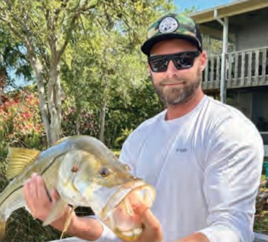 Capt. Brian Revak in a camo cap and sunglasses holding up a snook in front of a house and trees