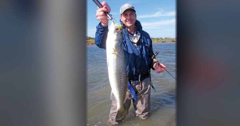 A smiling angler in a cap and blue rain jacket wading in the shallows, holding up a big speckled trout on a fish grip in one hand and his rod in the other