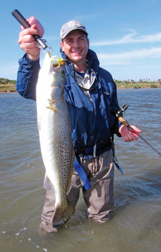 A smiling angler in a cap and blue rain jacket wading in the shallows, holding up a big speckled trout on a fish grip in one hand and his rod in the other