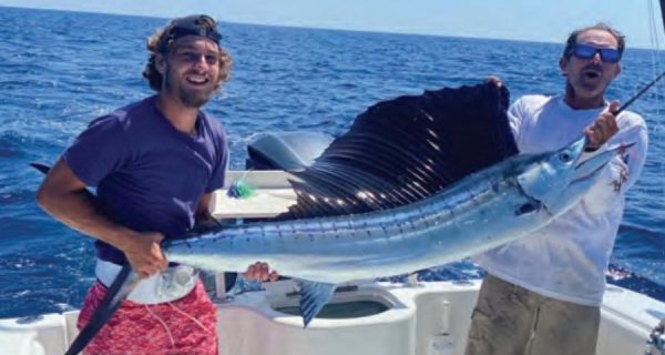 Two anglers on a boat offshore holding up a sailfish with its dark sail raised, one in a backward cap and purple shirt and the other in blue mirrored sunglasses