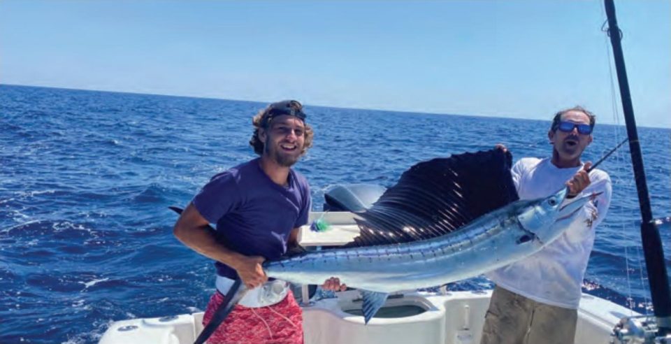 Two anglers on a boat offshore holding up a sailfish with its dark sail raised, one in a backward cap and purple shirt and the other in blue mirrored sunglasses