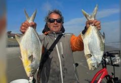 Marco Pompano in sunglasses and a gray and orange jacket holding up a big pompano in each hand on the beach, with a red bicycle beside him