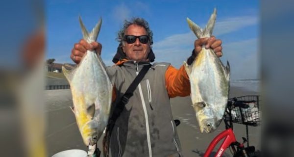 Marco Pompano in sunglasses and a gray and orange jacket holding up a big pompano in each hand on the beach, with a red bicycle beside him