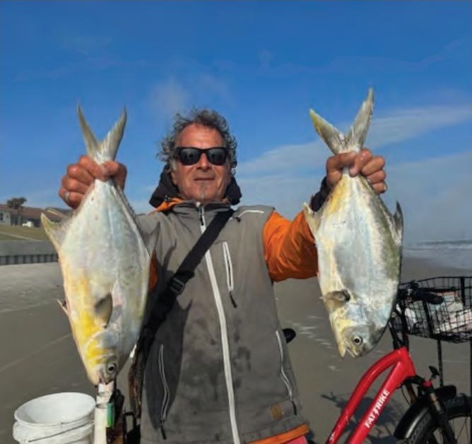 Marco Pompano in sunglasses and a gray and orange jacket holding up a big pompano in each hand on the beach, with a red bicycle beside him