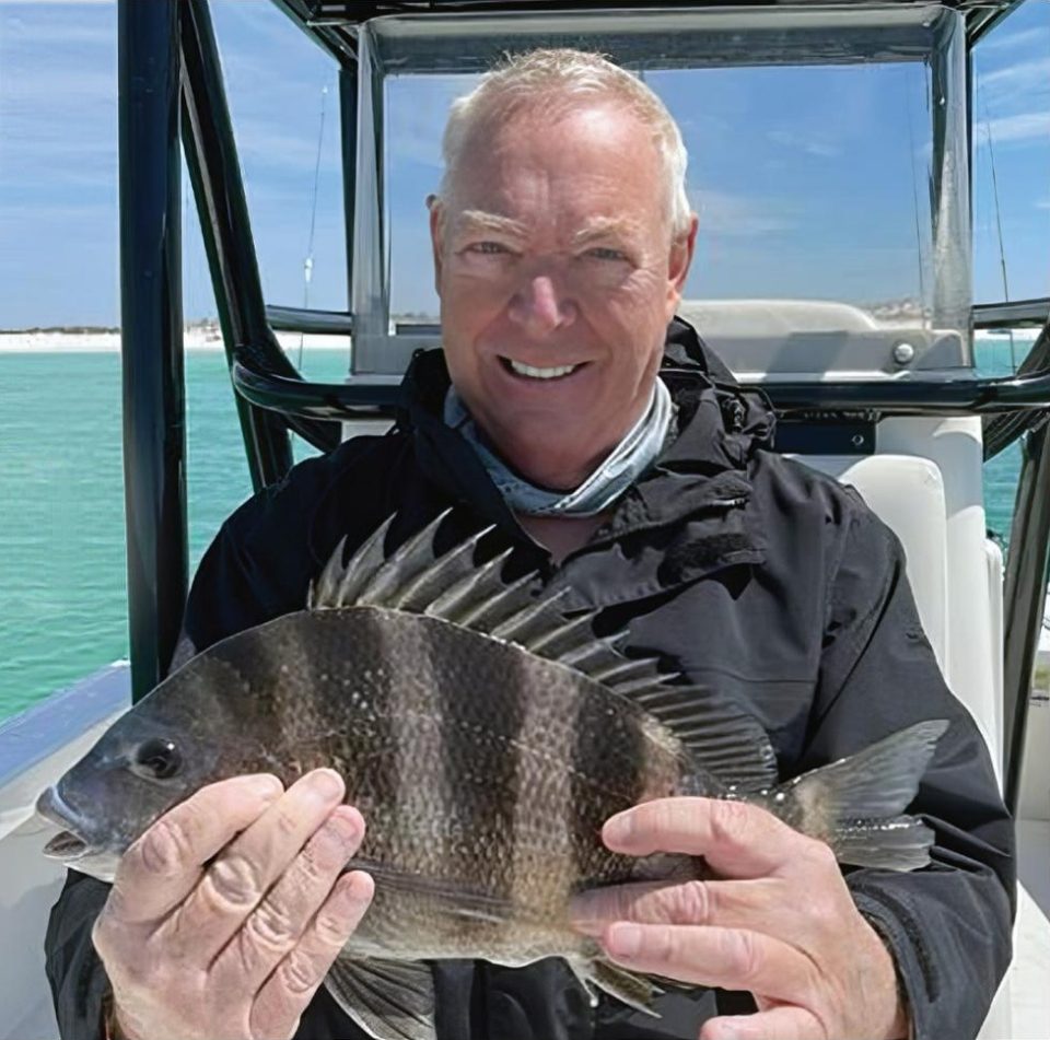 Gregg Lewis smiling on a boat under a T-top, holding up a sheepshead with turquoise water and a white sand beach behind him