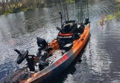 An orange and black Old Town Autopilot fishing kayak rigged with electronics and rods, floating on dark water with trees behind it