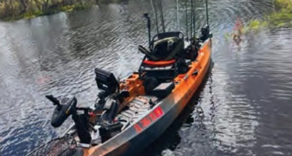 An orange and black Old Town Autopilot fishing kayak rigged with electronics and rods, floating on dark water with trees behind it