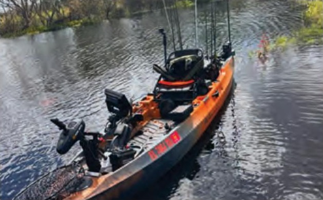 An orange and black Old Town Autopilot fishing kayak rigged with electronics and rods, floating on dark water with trees behind it