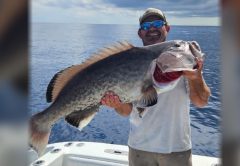 Capt. Chris Endicott in a cap and blue mirrored sunglasses holding up a huge grouper on the deck of his boat, with the open ocean behind him